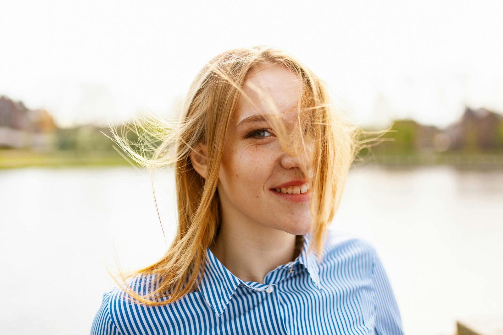 Portrait of a smiling woman with freckled face and wind-blown hair in a sunny outdoor setting.